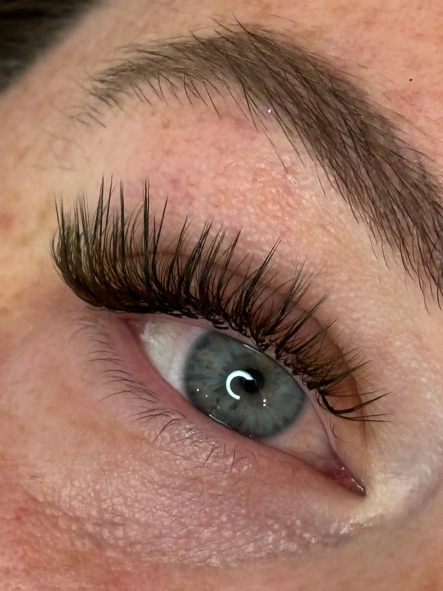 The right eye of a woman with blue eyes and light brown eyebrows. The eyelashes have extensions on them. The natural lash is thick and healthy.