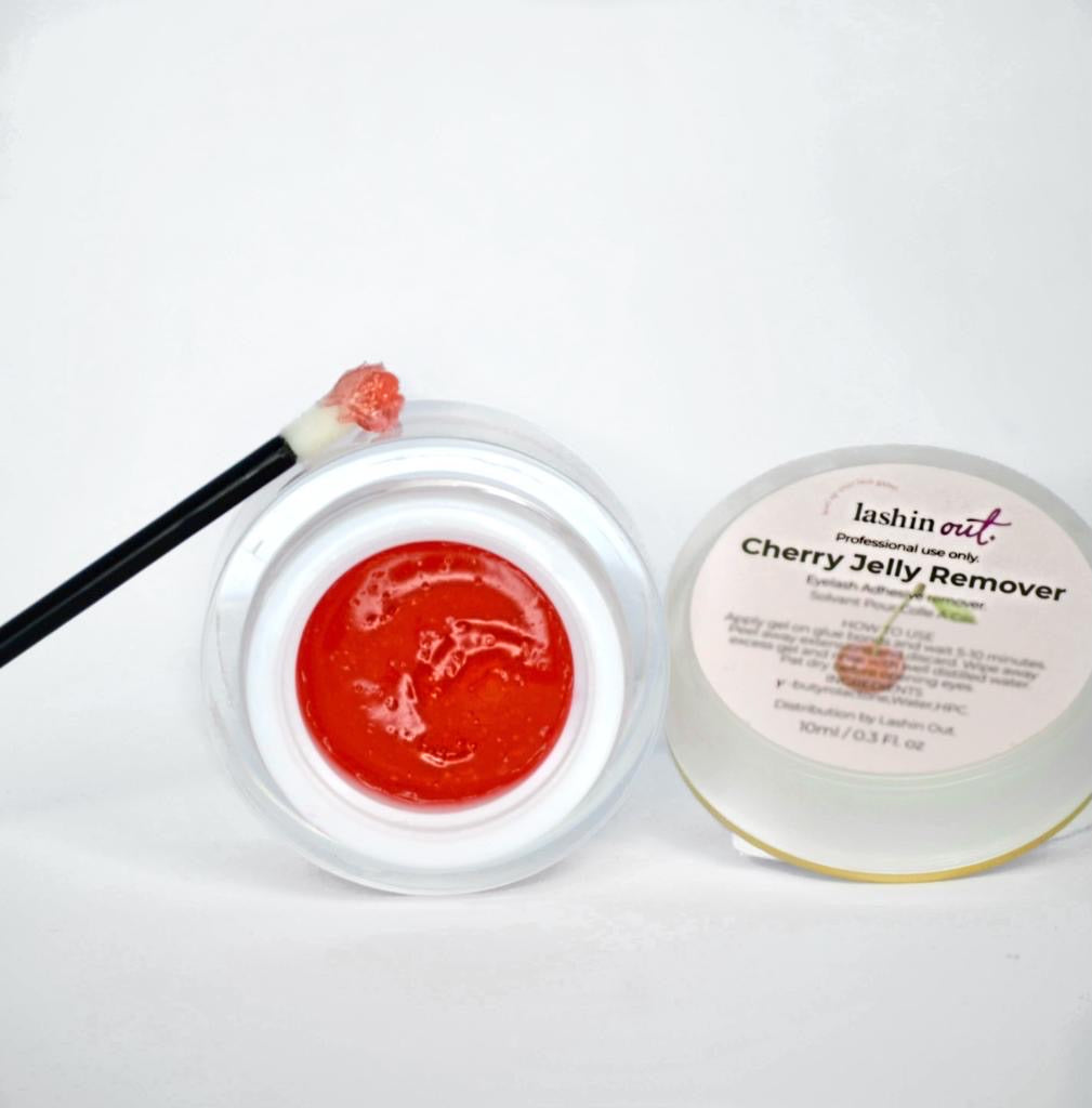 A round container with a label that says Lashin Out Cherry Jelly Remover, eyelash adhesive remover. The jar is opened, revealing a red jelly substance. There is a brush leaning against the jar with a bit of the red jelly remover on the brush. 