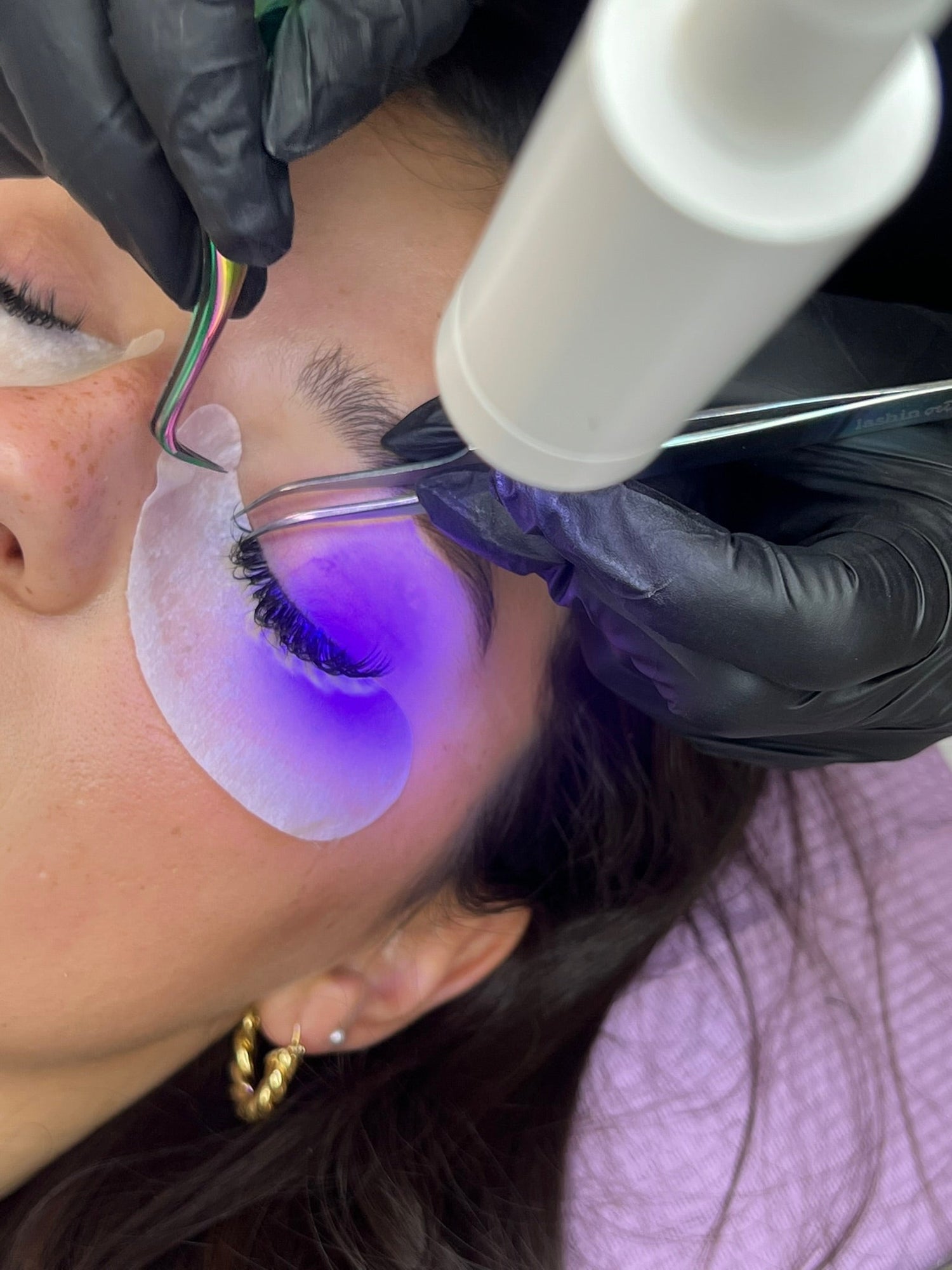 Close-up of a lash artist using tweezers and a UV LED light to cure eyelash extensions during application, with protective eye pad beneath the lashes.