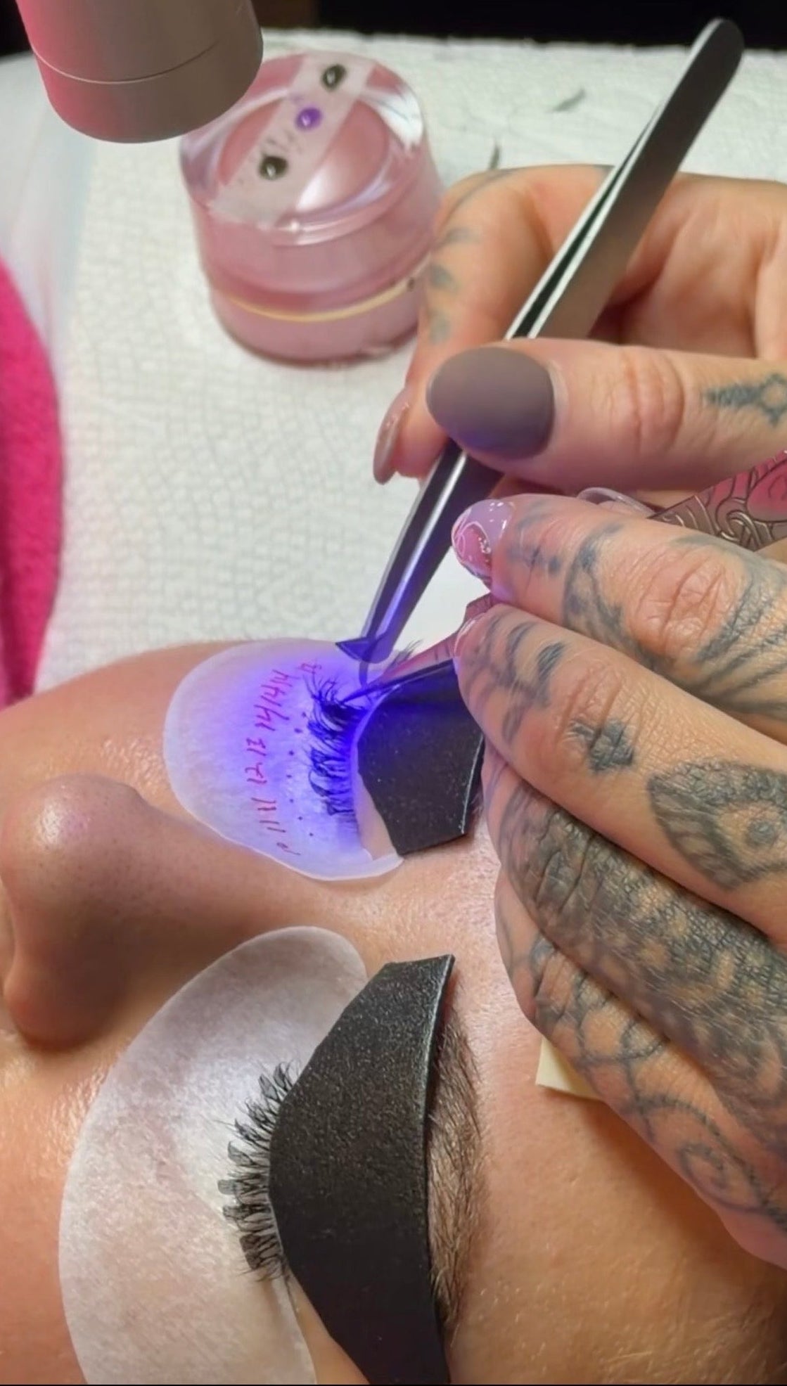 Close-up of a lash technician applying UV lash extensions with tweezers under a blue curing light, with under-eye pads and protective shields in place