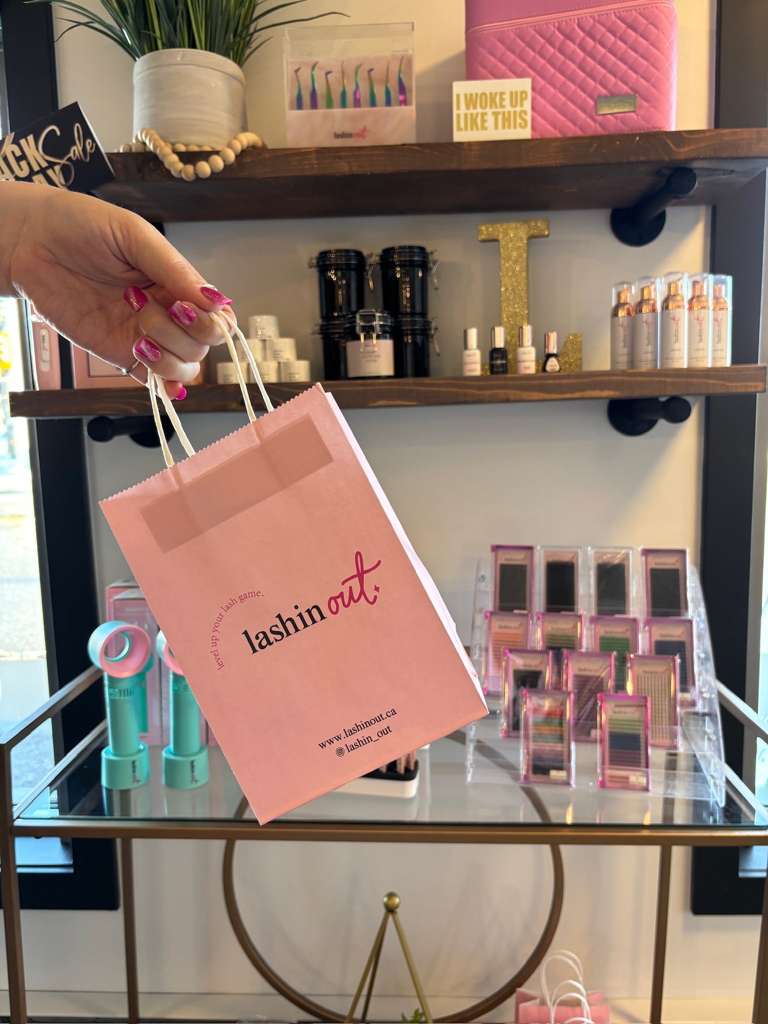 A women is holding a pink paper bag with a white handle. The bag says "lashin out". Behind the bag is a shelf with Lashin Out lashing products. There is Lash Suds lash cleanser, eyelash extension glues, and different trays of eyelash extensions. 