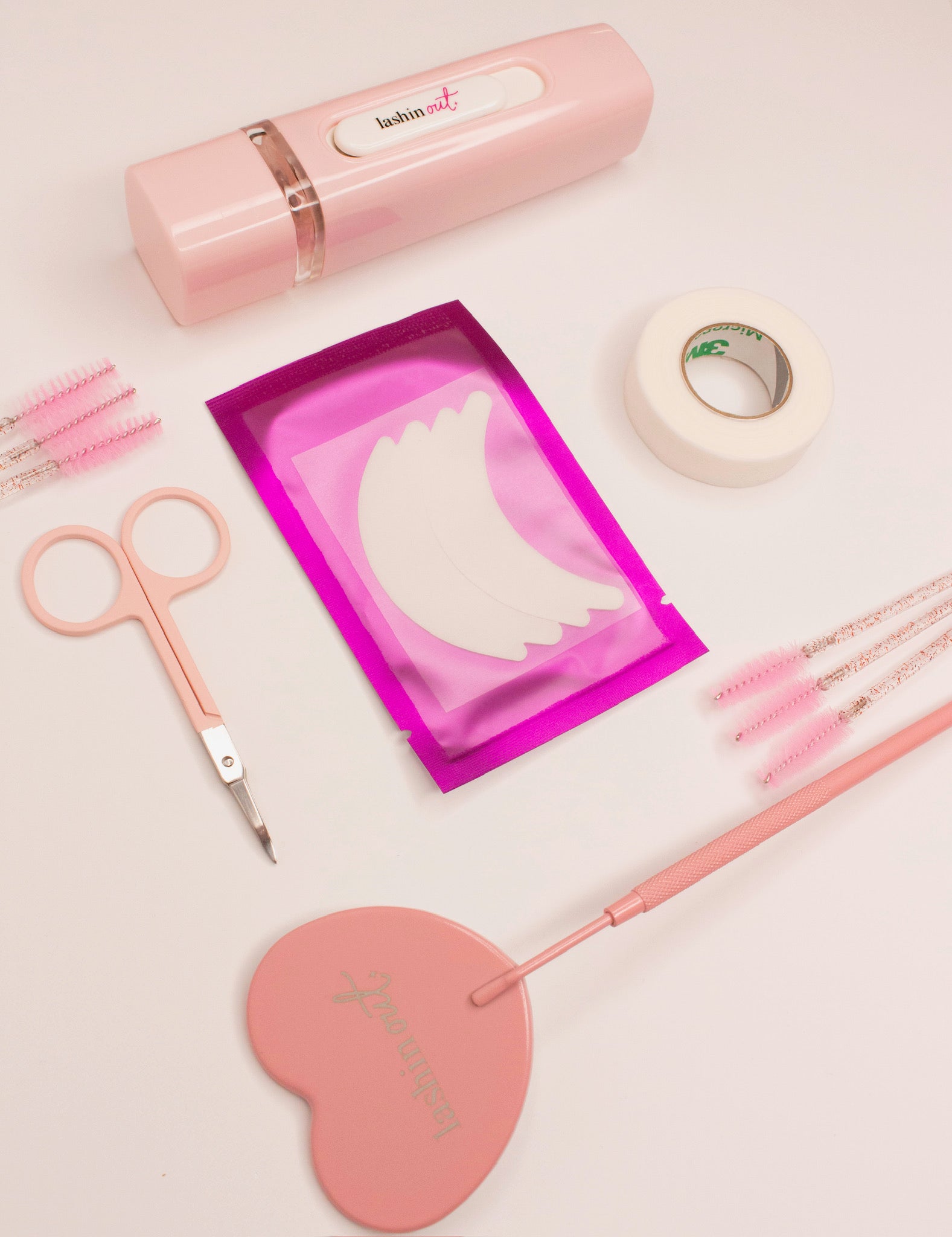 A variety of lash extension products are laid out. A small pair of pink scissors, 6 spoolies, a heart shaped mirror, a nano mister, surgical tape and hydrogel eyepads. 