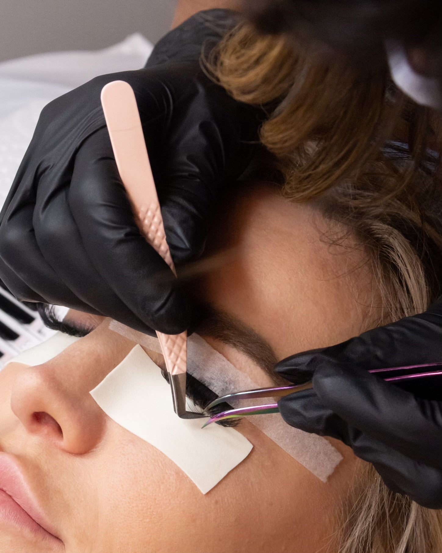 A woman is laying down with her eyes closed getting eyelash extensions applied. The lash artist is wearing black latex gloves and is holding pink tweezers in both hands. The last artist is applying an eyelash extension to the woman's left eye. 