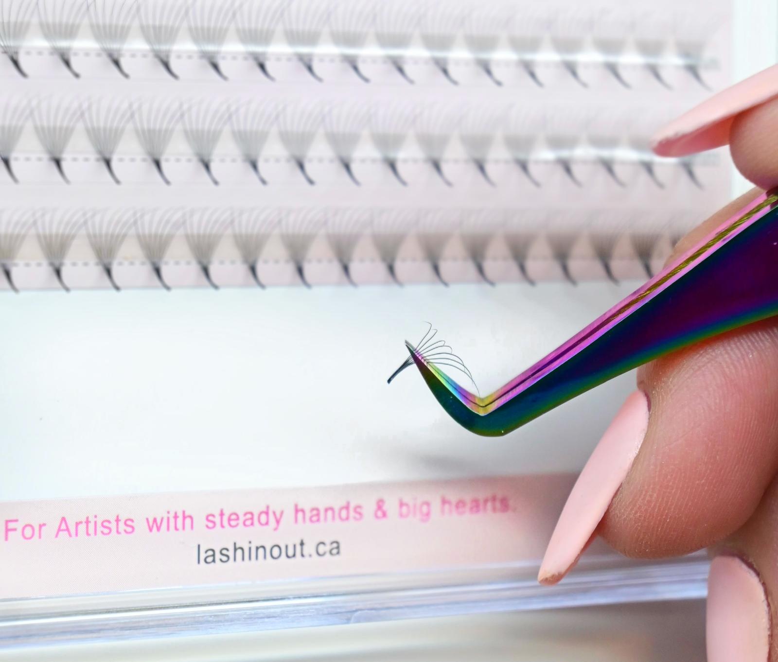 A lash artist is holding tweezers with a premade lash fan in the tweezer. In the background is a tray of premade lash fans. At the bottom of the tray it says "For artists with steady hands & big hearts."