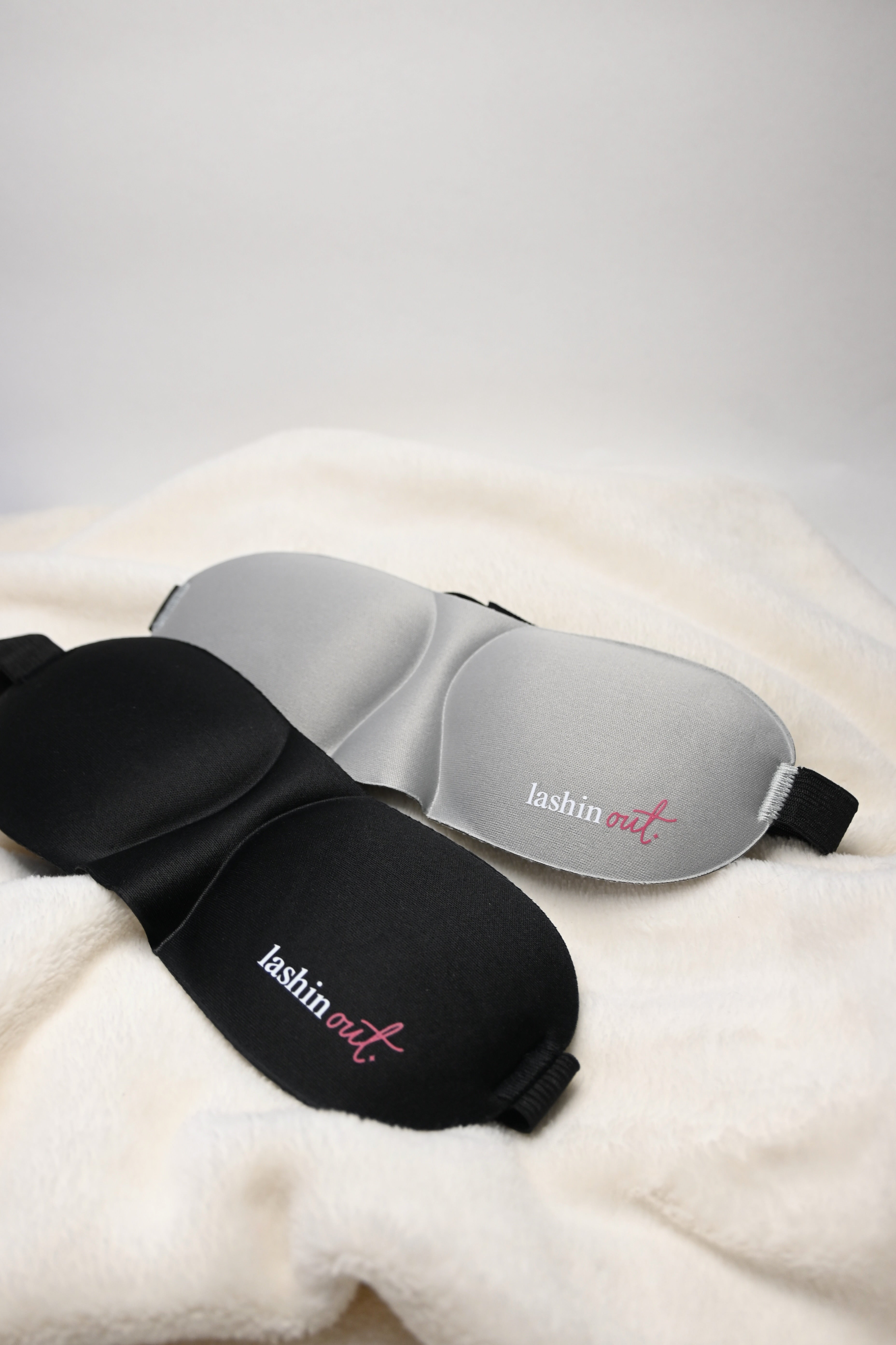 One black eyelash extension sleep mask and one grey eyelash extension sleep mask. Lashin Out is written in white and pink lettering on one eye cup.